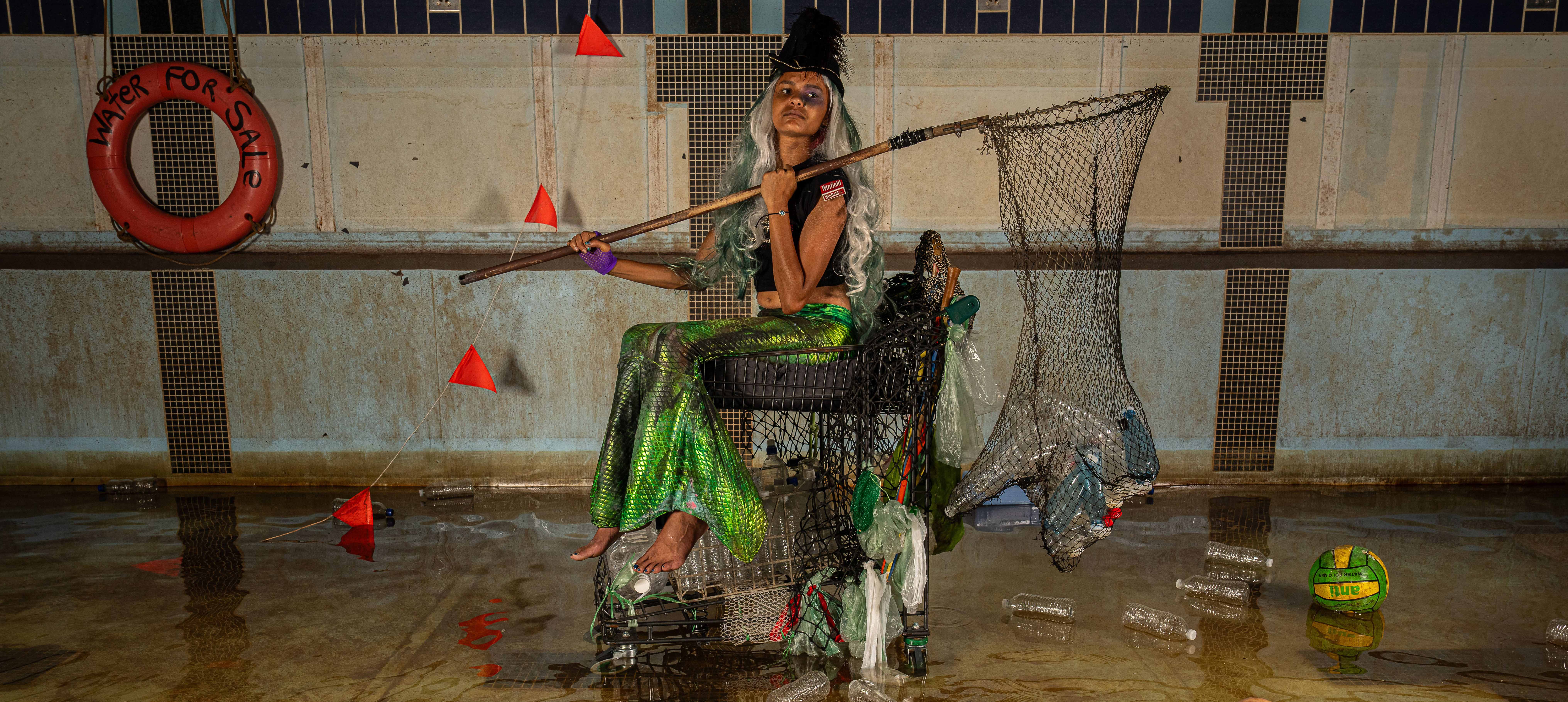 A photograph of a young girl, dressed as a mermaid. Her outfit looks to be made from found or discarded objects. She sits in a shopping trolley with a net over her shoulder which is filled with trash. The trolley is in a near empty pool. Around her is green water and floating debris. 