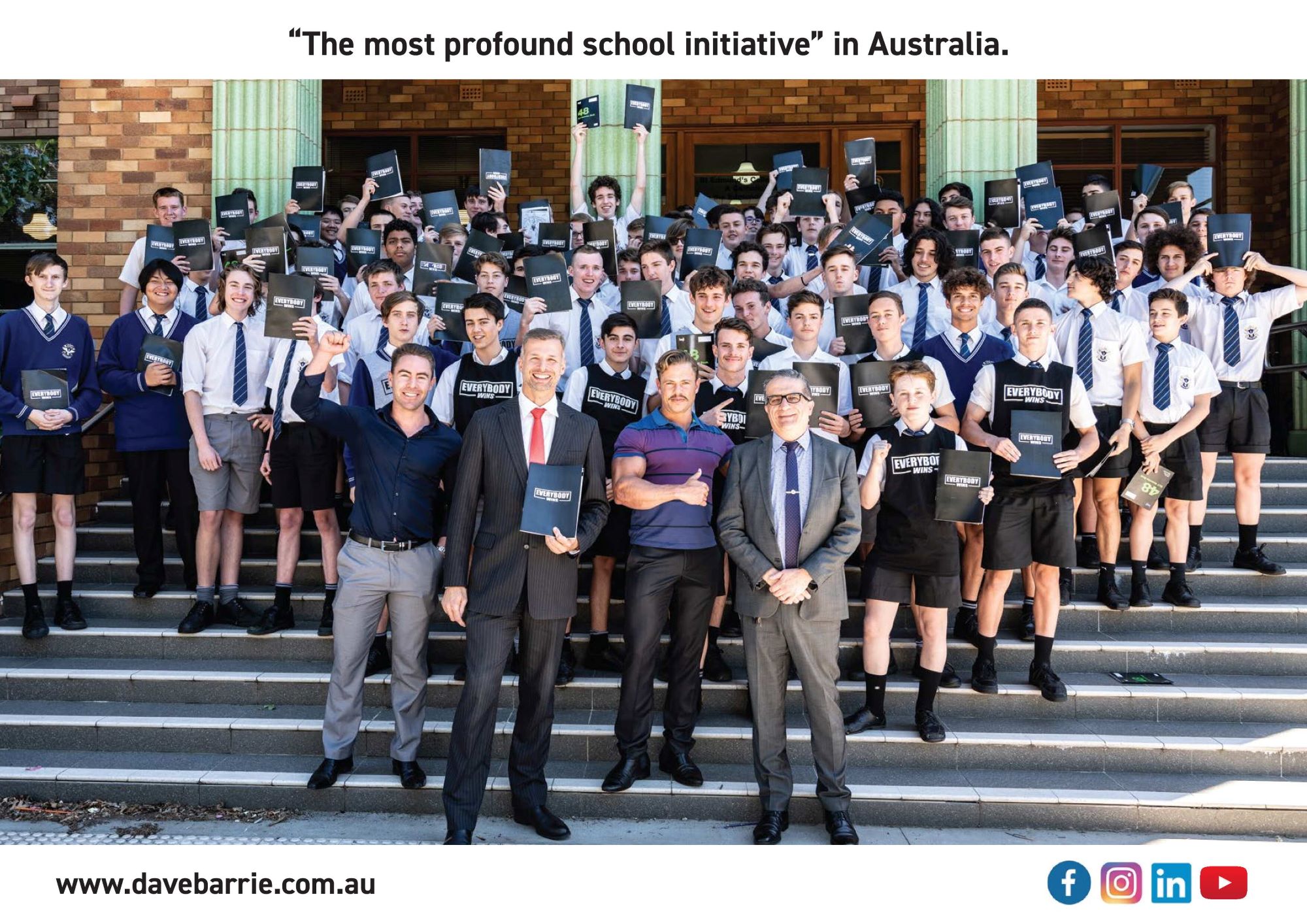 Dave Barrie standing on school steps, teachers to the right and students lined up on the stairs behind him holding booklets that say Everybody Wins
