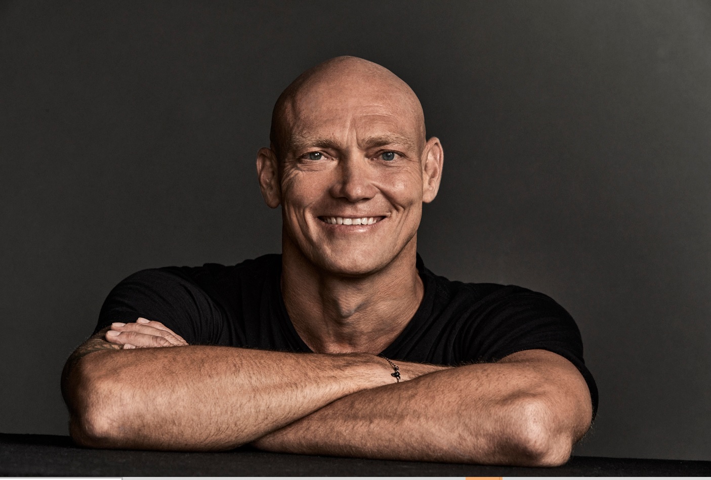 A bald man with a wide smile, dressed in black, looks out across this folded arms