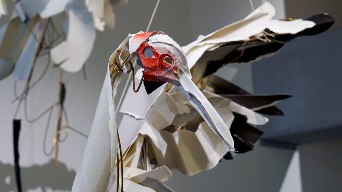 A photograph of a brolga sculptures head close up. Red, white and brown papers form the surface while we can see the brass skeleton through the gaps between materials. 