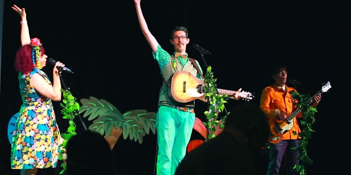 One character in colourful dress with microphone and two characters with musical instruments holding up their arms