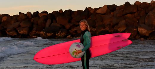 A photograph of a young female surfer, under one arm is a pink surfboard, in her other hand, is a globe. The picture is taken at sunset so the image has the warm glow of twilight. 