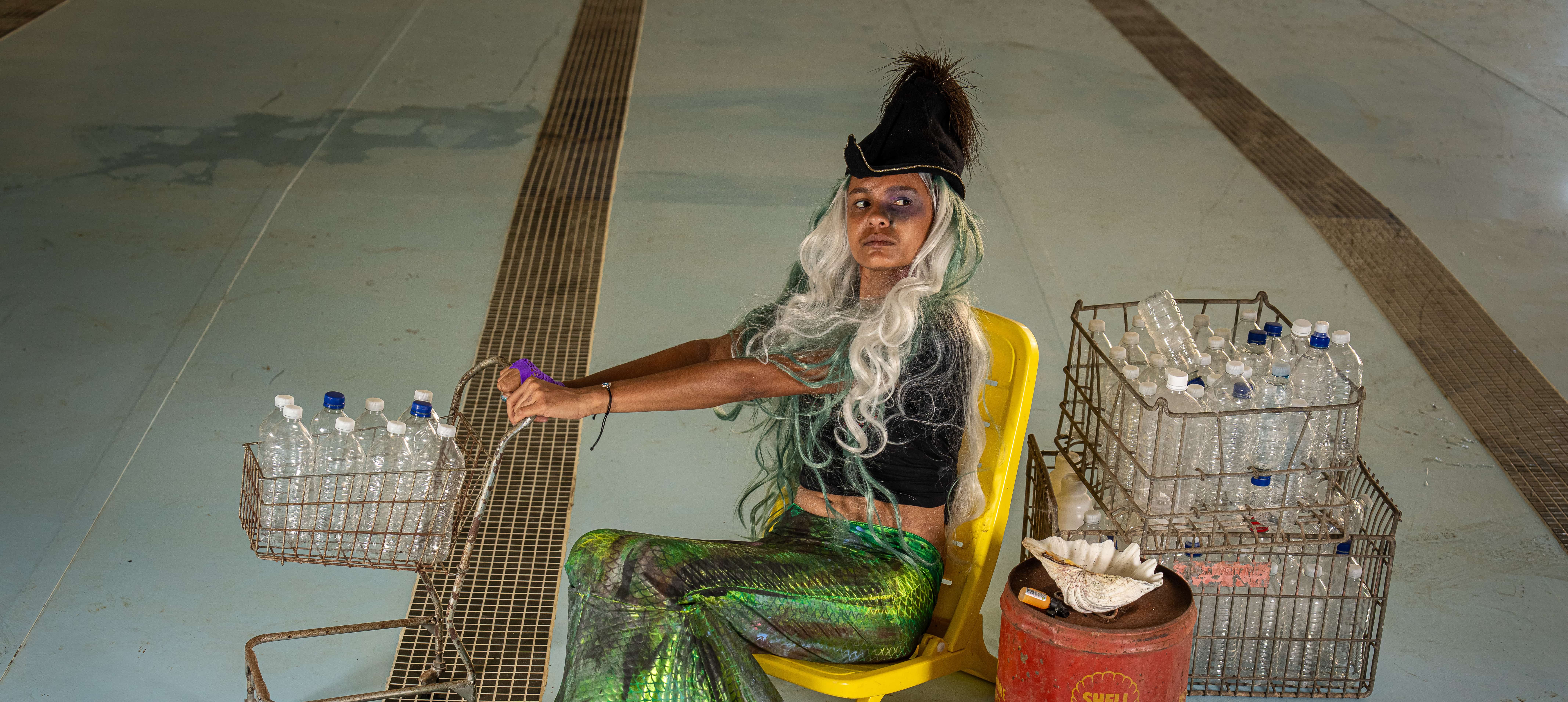 A photograph of a young indigenous girl in a green mermaid dress, she is seated on a yellow chair and holds a toy shopping trolly filled with plastic bottles. Behind her are baskets of bottles, a rusted petrol can and a shell. She wears a hat and a wig with long blonde hair. The arrangement is on the bottom of an empty pooll