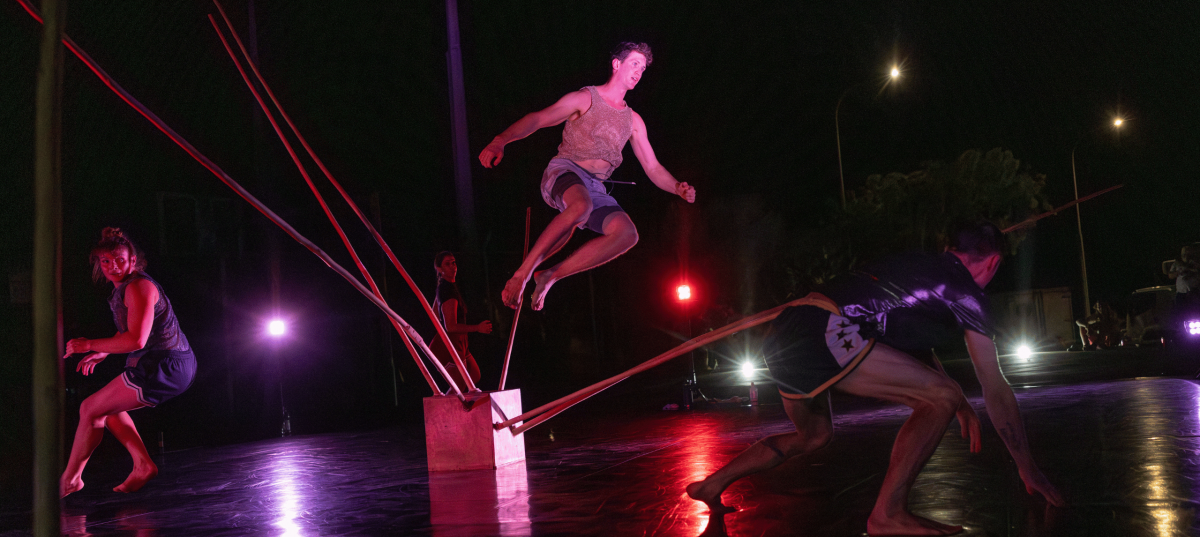 Three dancers leaping above near an acrobatic rig, night performance surrounded by red light