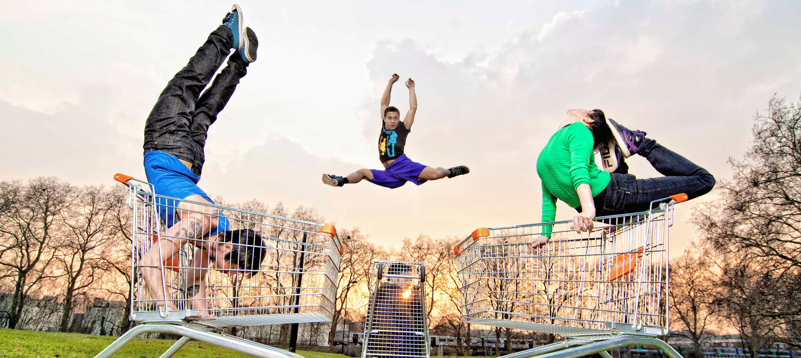 Three dancers in an outdoor space, two dancing on shopping trolleys, one dancer is leaping in the air behind them.