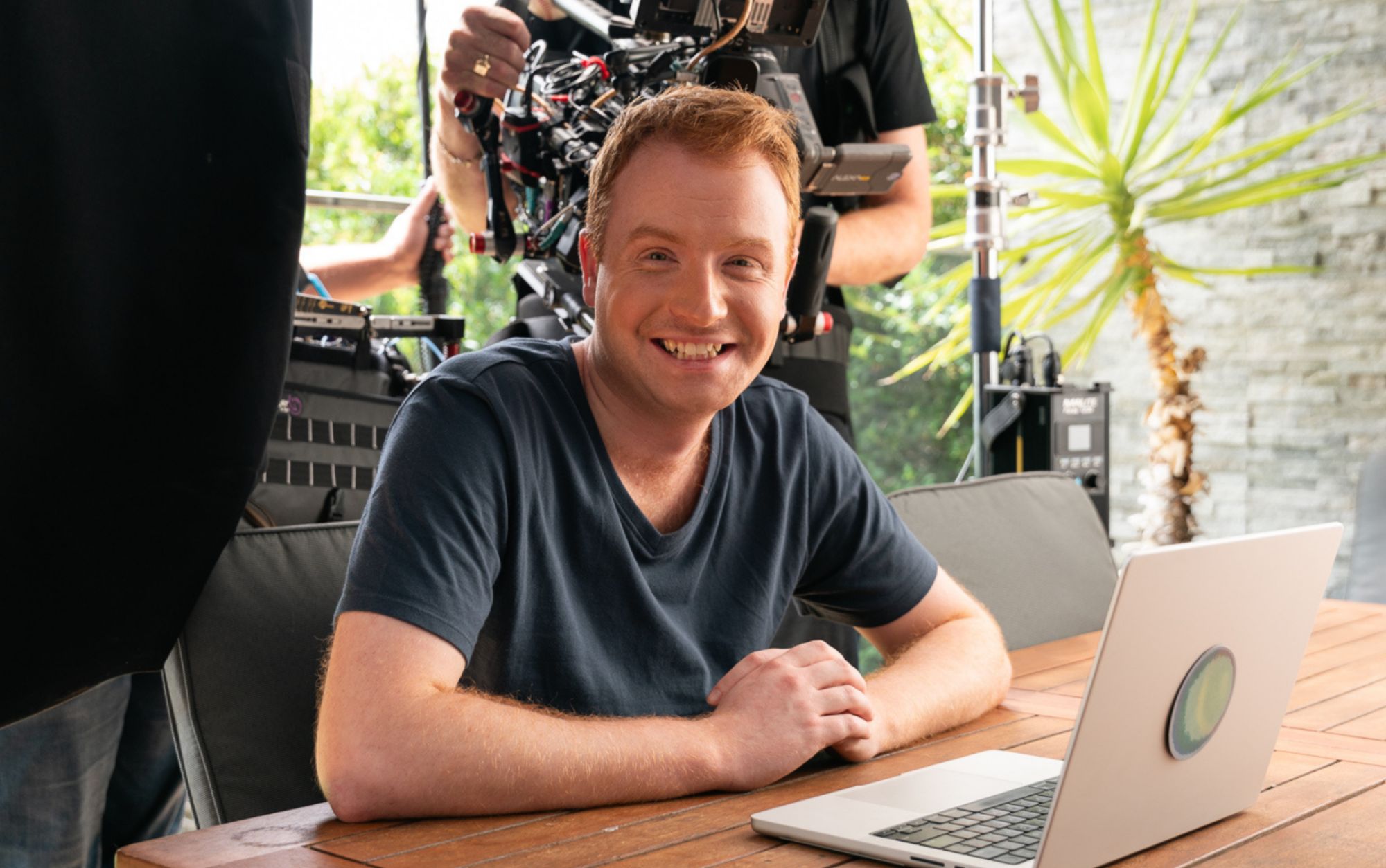 Young man smiling at camera in front of laptop with large tv camera filming behind him