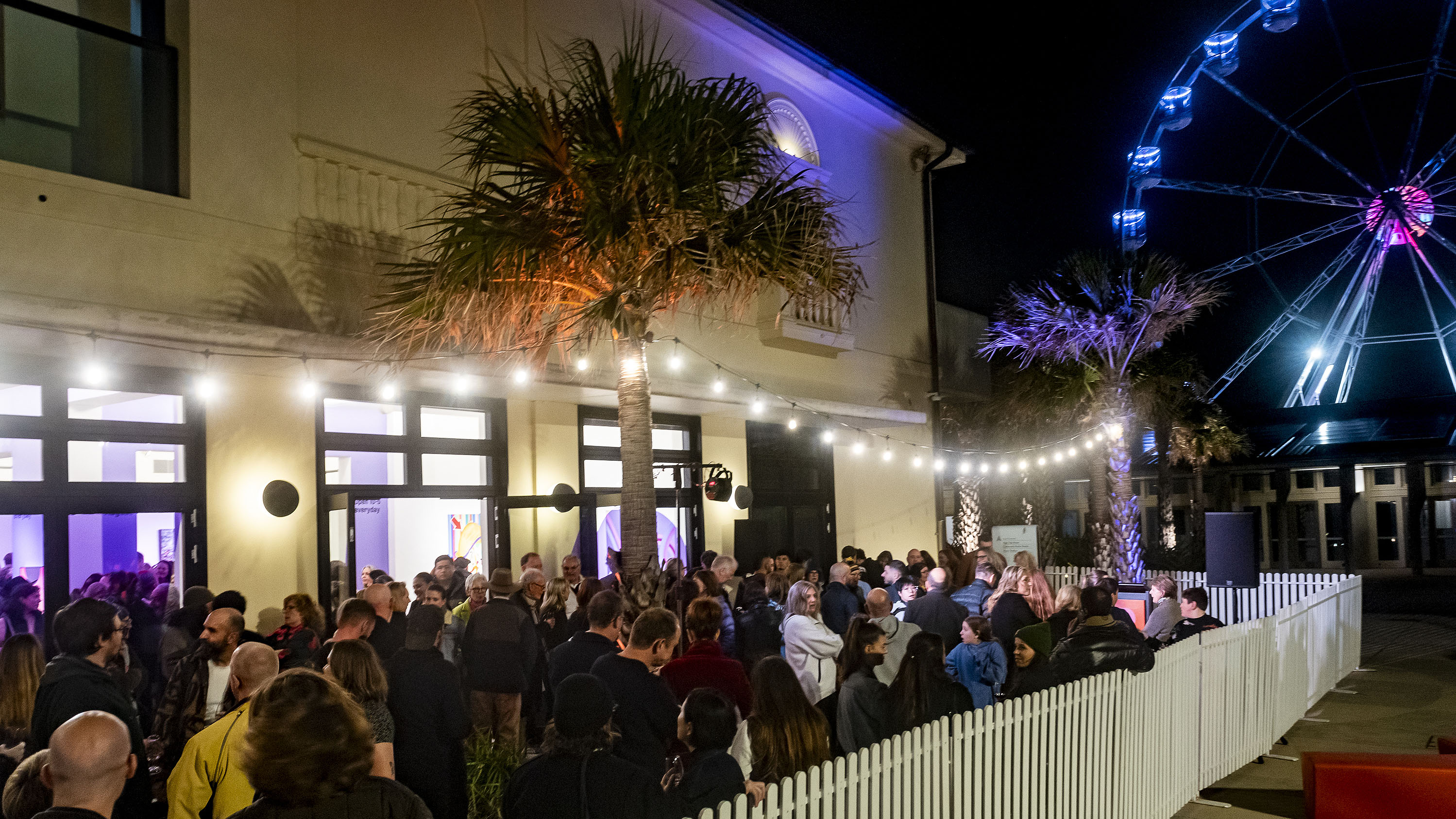 A photograph of a crowd assembled outside the Bondi Pavilion Gallery at night. Behind them, an illuminated Ferris wheel can be seen.