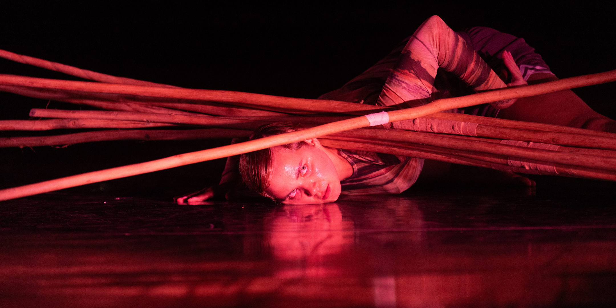 A dancer lying on floor with their head surrounded by large sticks laying parallel to head, night stage with red lights