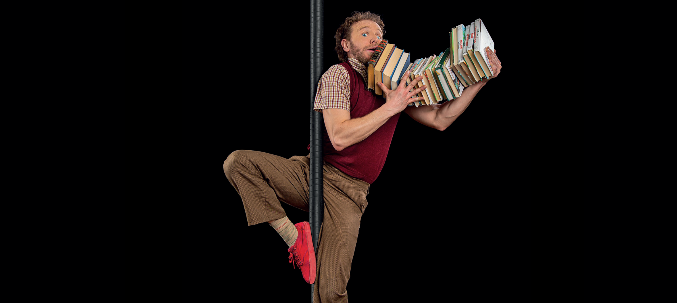 The actor with brown clothes and red shoes is hanging off a pole with a bunch of books that are just about to fall out of his hands.