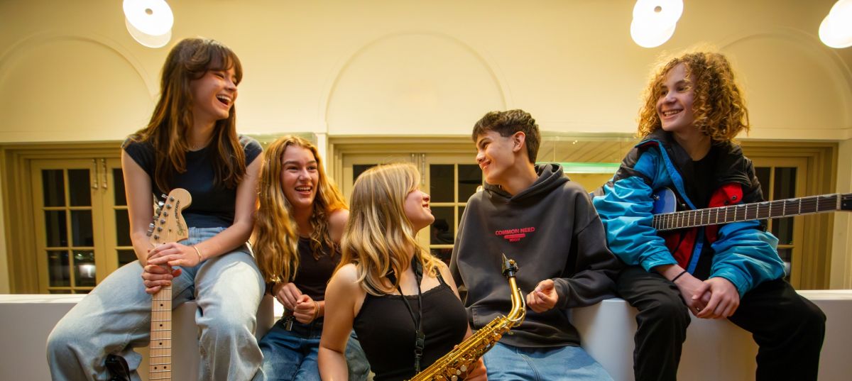 A Band of boys and girls laughing with some instruments and sitting on a bar