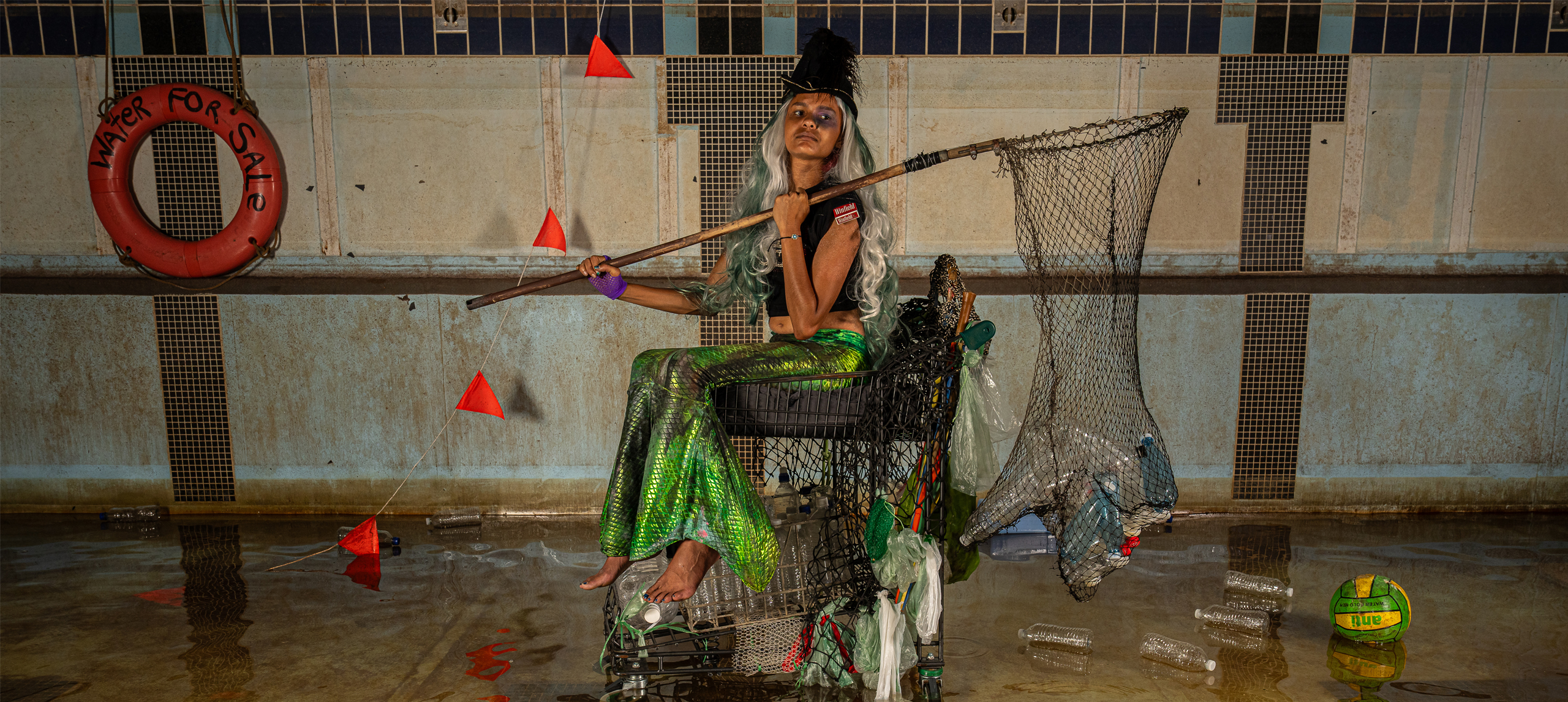 A photograph of a girl in mermaid outfit in a semi-flooded pool. She is dressed as a mermaid in second hand objects and recycled materials, she holds a new filled with trash and around her is floating debris. 