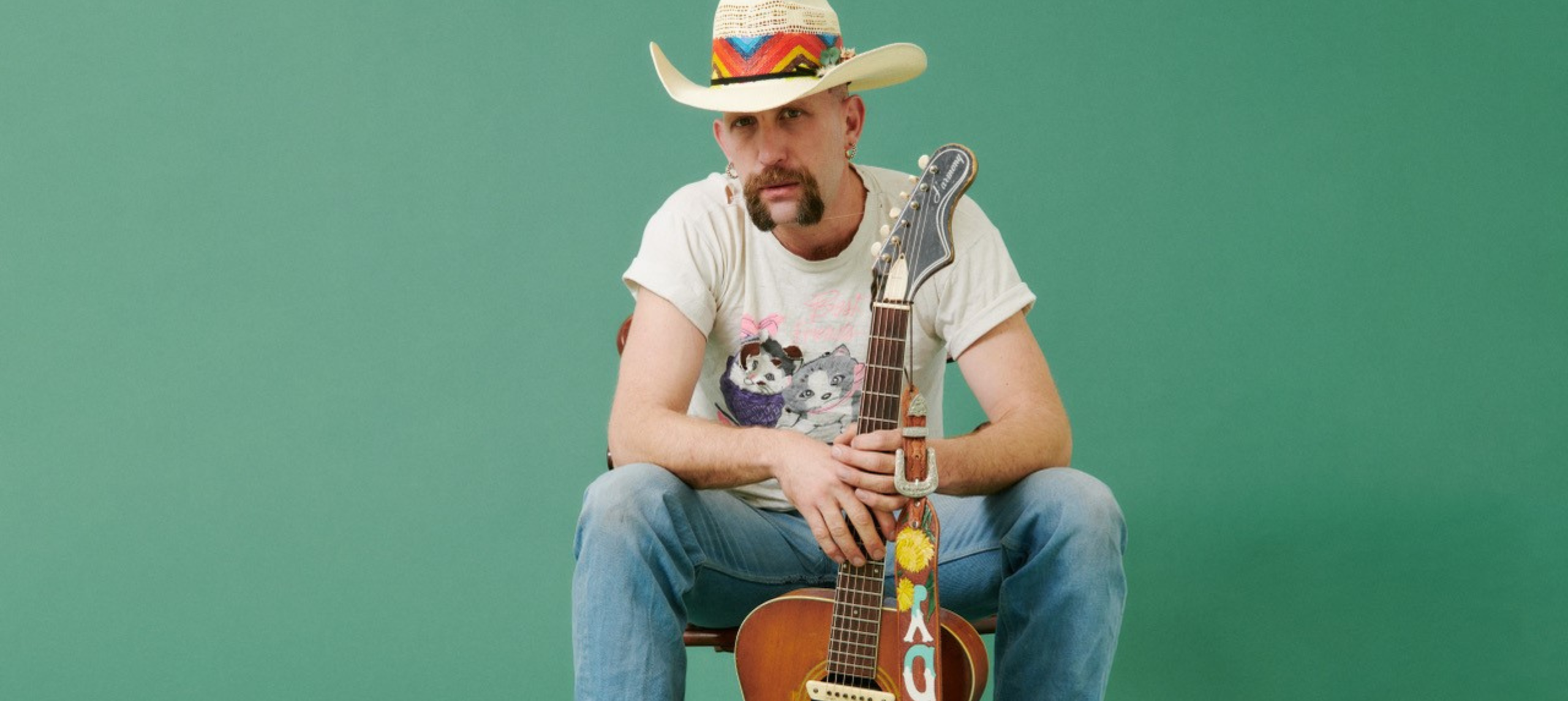 A country music artist with a big hat sitting on a chair with his guitar