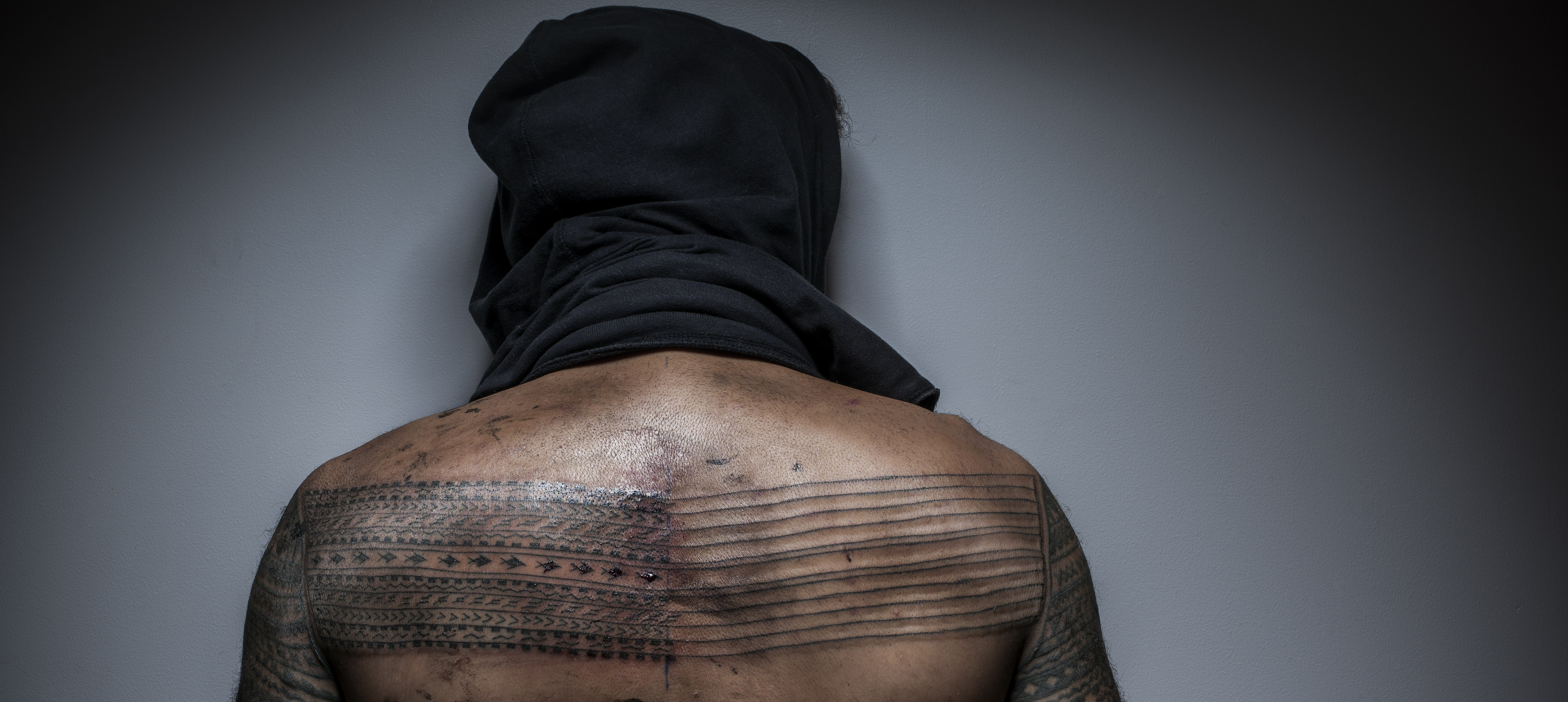 The artists bare back is presented to us, his head is under a cloth. Across his back are a series of Samoan tatoos running in horizontal lines across his flesh.