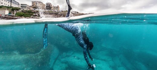 A photograph of a diver submerging, the bottom half of the image shows below the ocean with cerulean tones, a female diver in a wet suit and rocky forms on the bottom of the sea. The top half of the image is muted grey tones with houses of North Bondi visible in the distance.