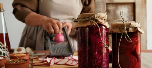 There are two full jars of pickled vegetables at the front of the picture, with a lady wearing an apron in the foreground who is using a mandolin to slide radishes onto a wooden cutting board. We is in the process of preserving vegetables.