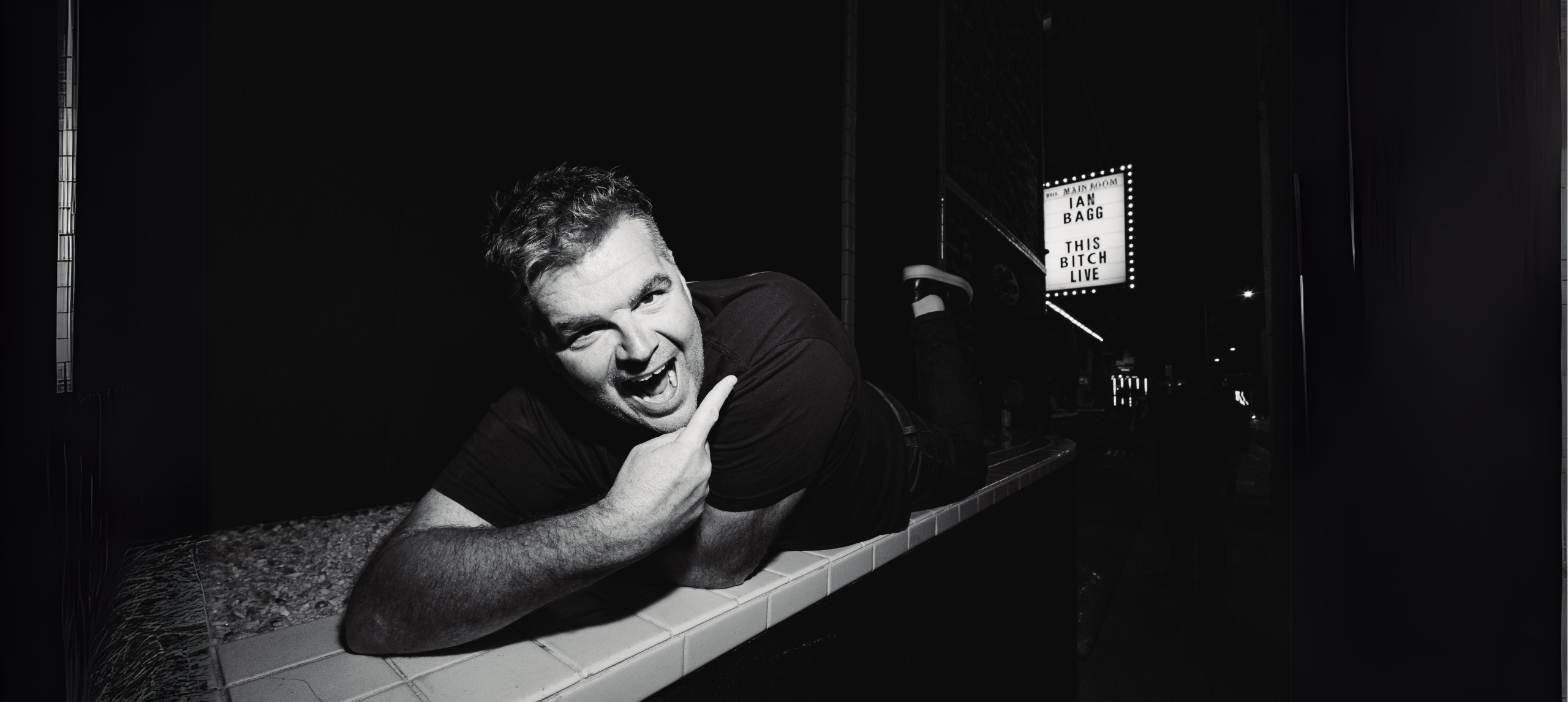 Ian Bagg comedian laying on ground pointing at marqui light with his name on it (black and white)